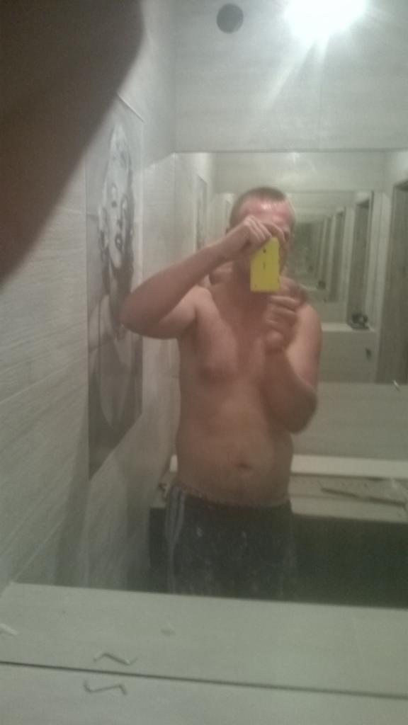 Mężczyzna bez koszulki robi sobie zdjęcie telefonem w łazience z lustrami i tapetą z Marilyn Monroe.