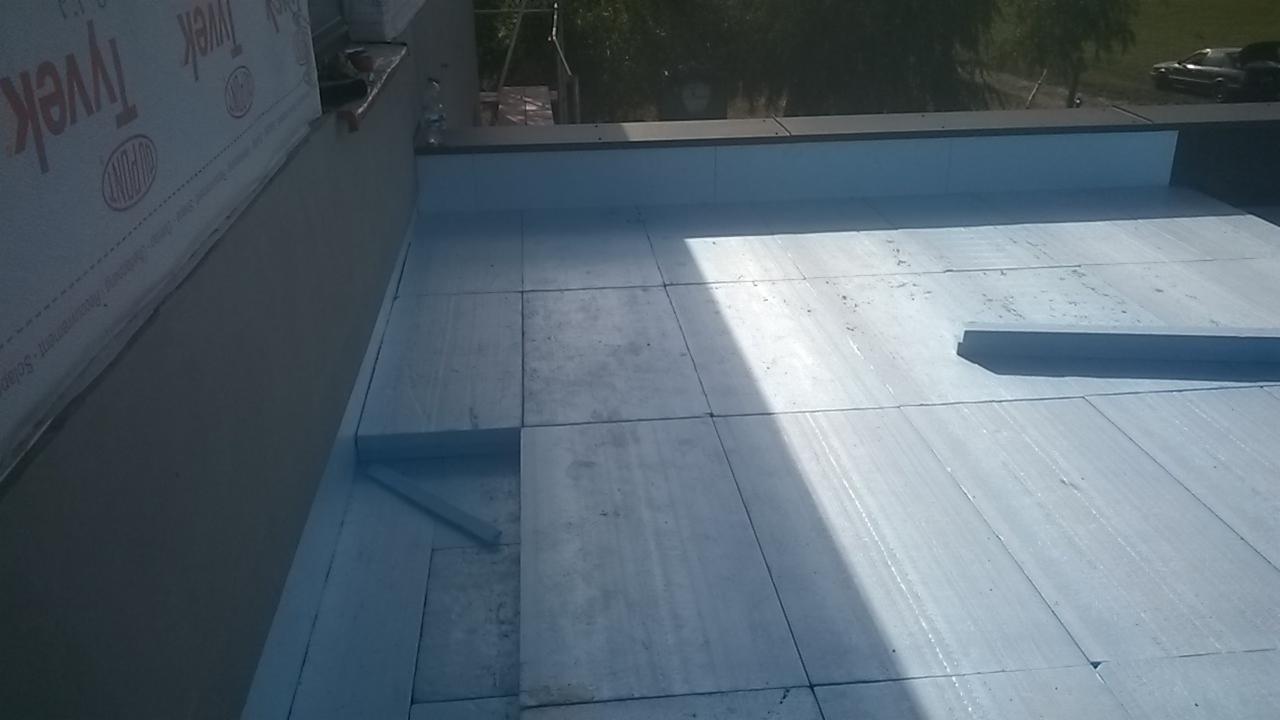 Płyty izolacyjne z polistyrenu ekstrudowanego (XPS) układane na płaskim dachu, widoczna membrana wiatroizolacyjna Tyvek na ścianie attyki.