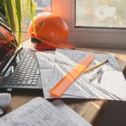 Biuro rzeczoznawcy budowlanego: Laptop, pomarańczowy kask ochronny, rysunki techniczne, linijka i cyrkiel na drewnianym biurku w pobliżu okna z rośliną doniczkową.