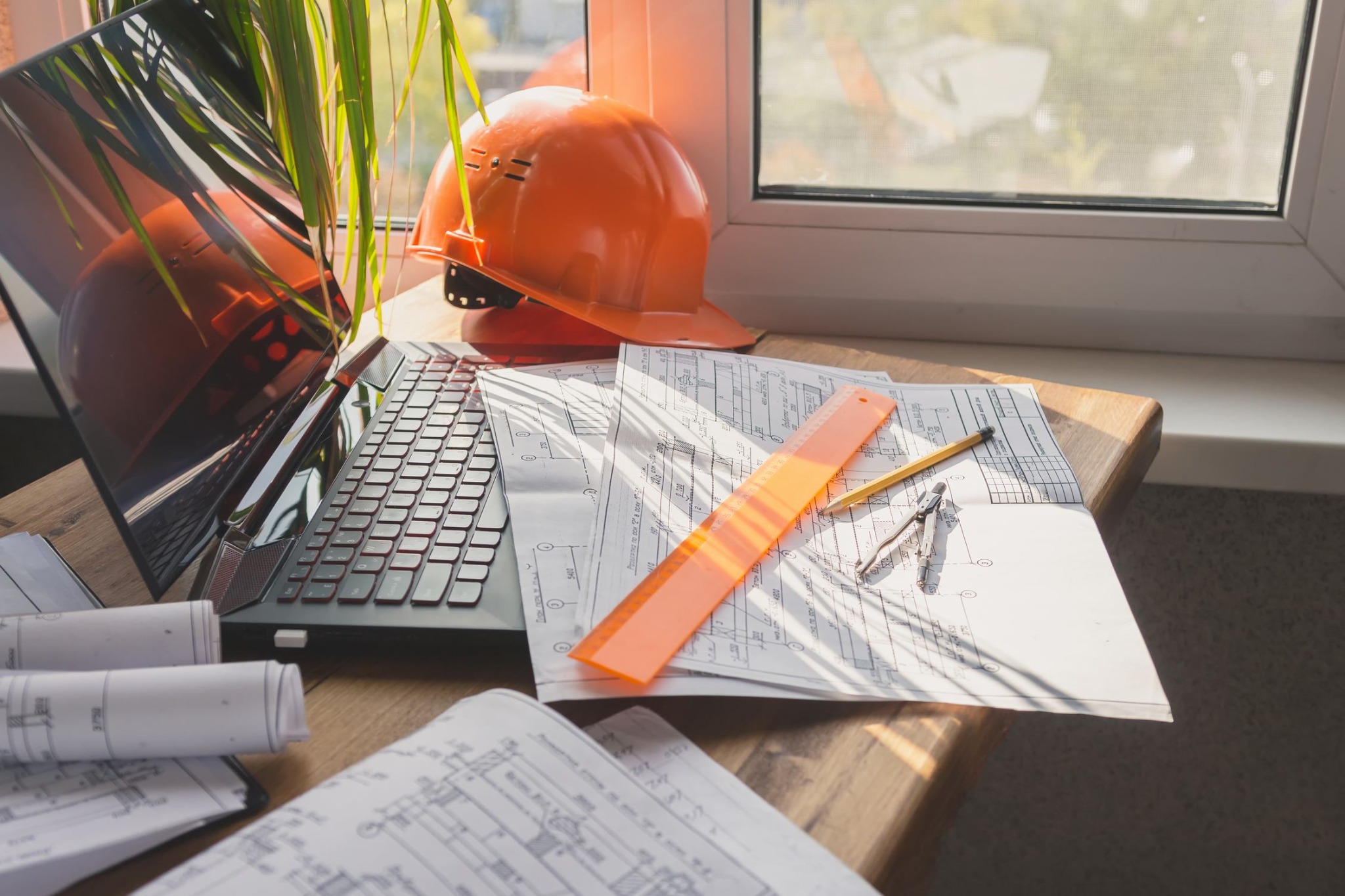 Biuro rzeczoznawcy budowlanego: Laptop, pomarańczowy kask ochronny, rysunki techniczne, linijka i cyrkiel na drewnianym biurku w pobliżu okna z rośliną doniczkową.