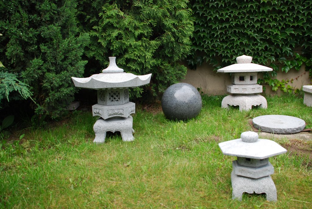 Ozdobne, kamienne lampiony ogrodowe i kula z granitu rozmieszczone w trawie, na tle zieleni krzewów i ściany porośniętej bluszczem.