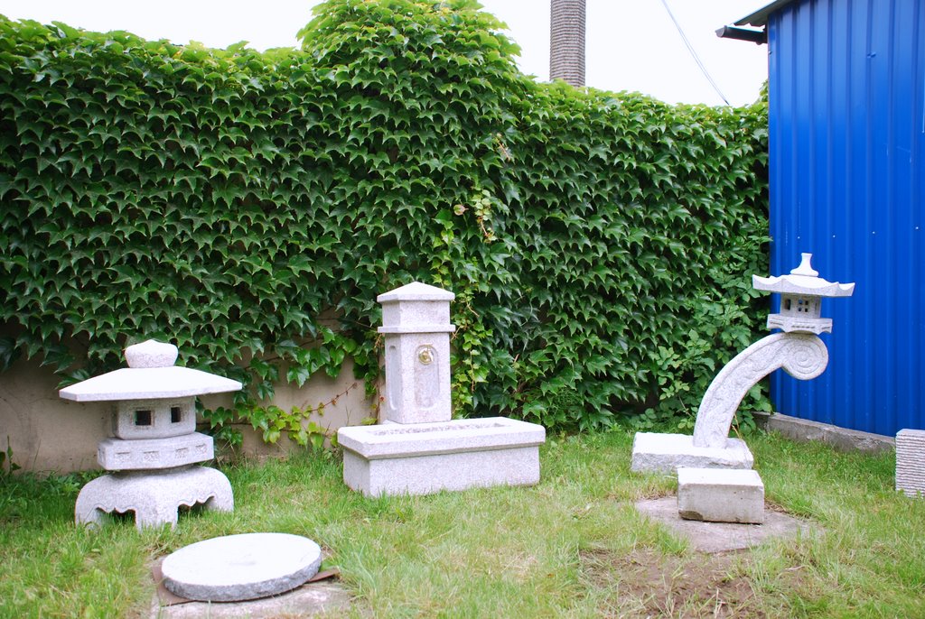 Dekoracyjne elementy z jasnego kamienia w ogrodzie: fontanna, dwie latarnie w stylu japońskim, tło stanowi ściana z gęstego bluszczu i niebieski blaszany budynek.