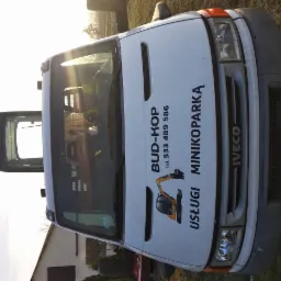 Przód białego samochodu dostawczego Iveco z nadrukowanym logo firmy BUD-KOP i numerem telefonu, zaparkowanego na trawie w słoneczny dzień.