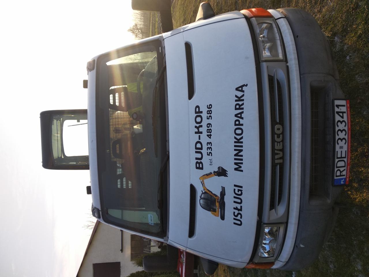 Przód białego samochodu dostawczego Iveco z nadrukowanym logo firmy BUD-KOP i numerem telefonu, zaparkowanego na trawie w słoneczny dzień.