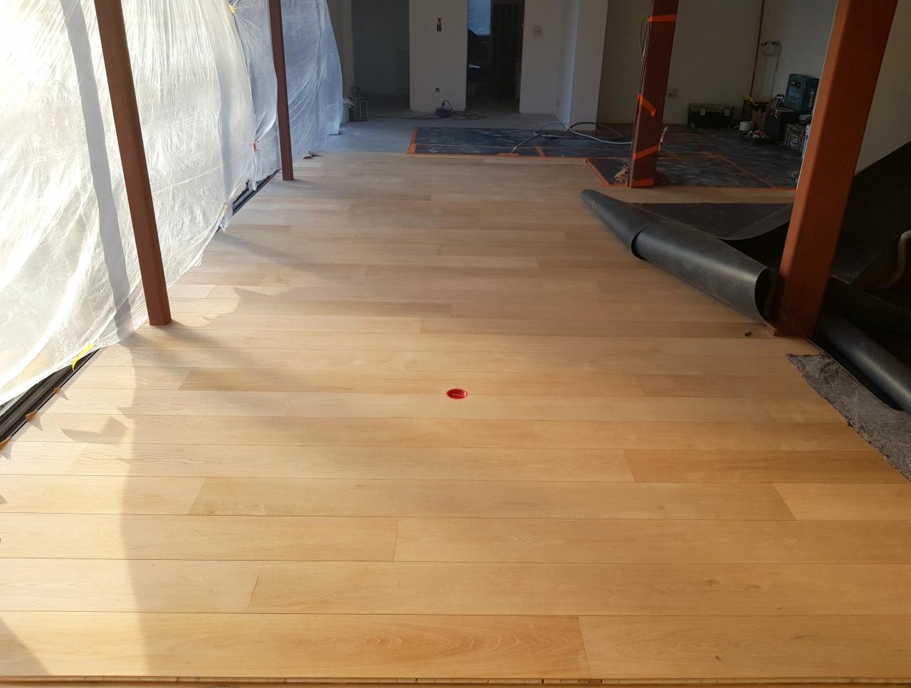 Świeżo położona, jasna podłoga drewniana w pomieszczeniu w trakcie remontu, z widoczną folią ochronną przy ścianie i elementami konstrukcyjnymi zabezpieczonymi pomarańczową taśmą.