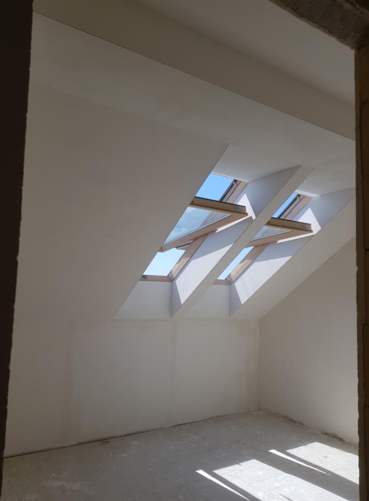 Poddasze po tynkowaniu maszynowym, widoczne dwa okna dachowe z drewnianymi ramami i fragment surowego muru w rogu pomieszczenia.