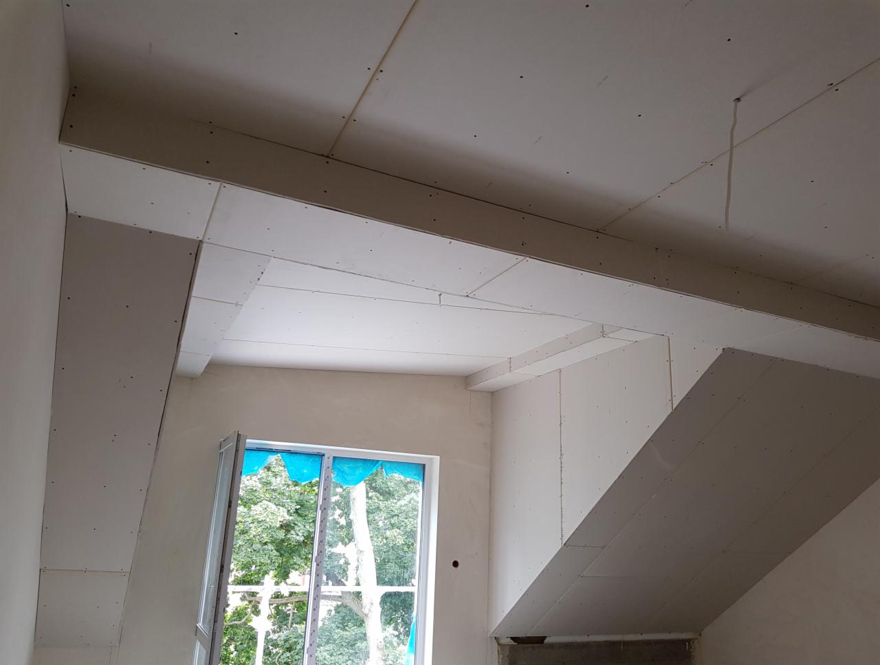Sufit podwieszany z płyt gipsowo-kartonowych w trakcie montażu, widoczne profile i łączenia, okno z zabezpieczeniem folią.