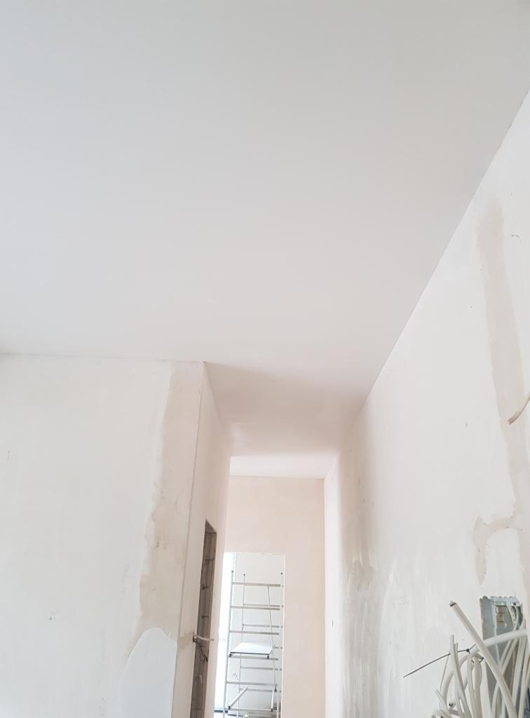 Świeżo położone, gładkie tynki na suficie i ścianach korytarza w trakcie remontu. Widoczne niedoskonałości i pozostałości po pracach wykończeniowych.