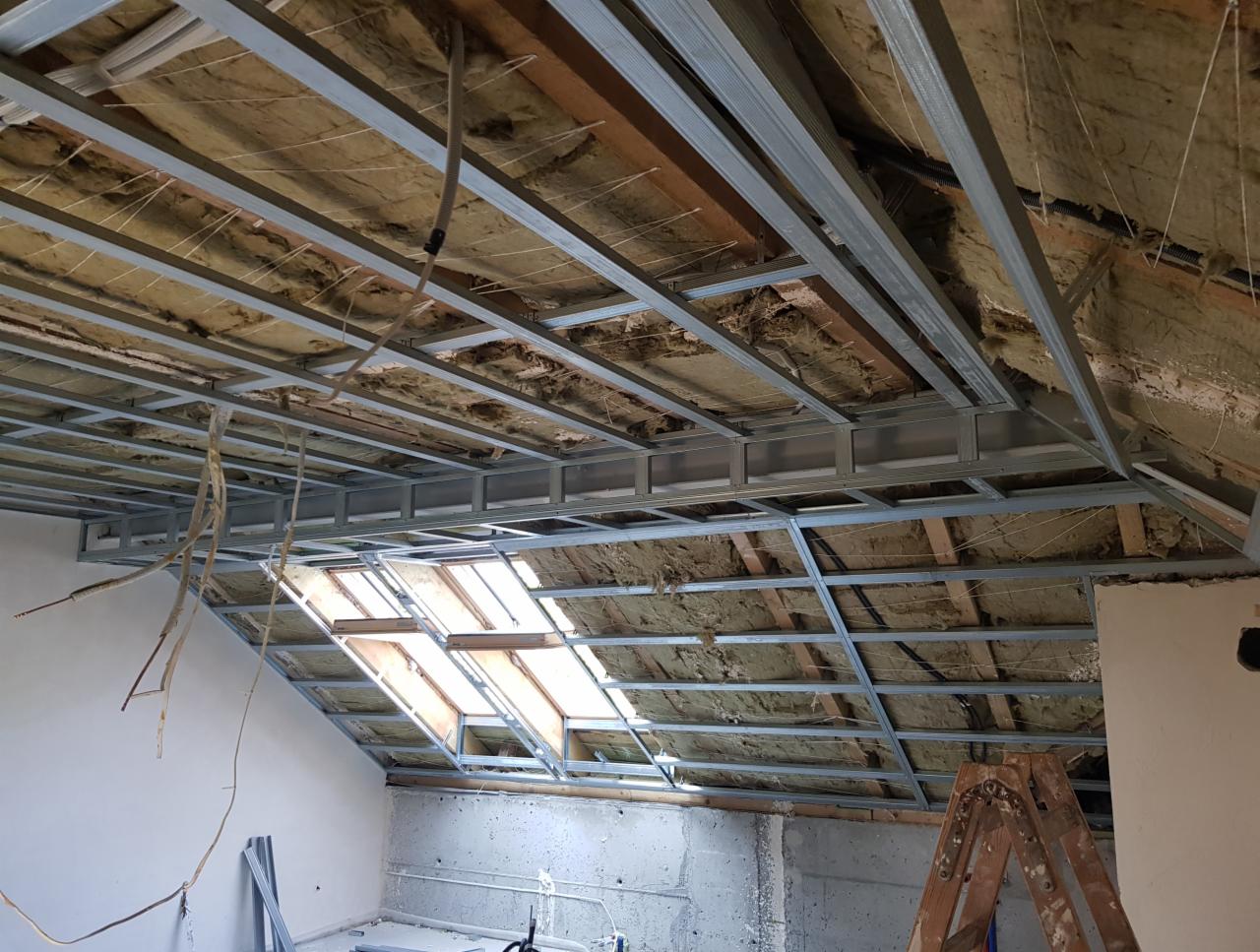 Montaż metalowej konstrukcji sufitu podwieszanego na poddaszu, widoczna izolacja z wełny mineralnej i okno dachowe, drabina oparta o ścianę.