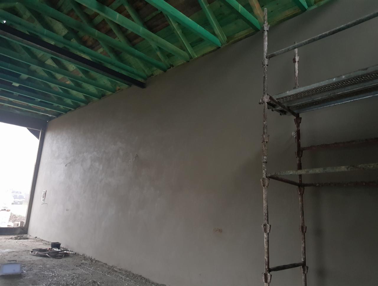 Świeżo nałożony, gładki tynk na ścianie wewnętrznej, widoczne rusztowanie i drewniana konstrukcja dachu, w tle otwór okienny.