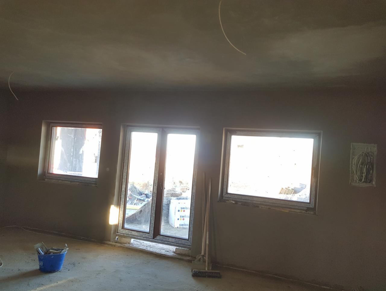 Świeżo nałożone tynki maszynowe na ścianach i suficie wnętrza, widoczne okna i drzwi balkonowe, niebieskie wiadro z narzędziami na podłodze, przewody elektryczne wystające ze ściany.