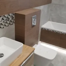 Nowoczesna łazienka z drewnopodobnymi i mozaikowymi akcentami: umywalka nablatowa, podwieszana toaleta z zabudową, wanna z podświetleniem LED, szare płytki.