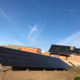 Panel słoneczny zamontowany na metalowej konstrukcji na tle budynków w trakcie budowy i błękitnego nieba z delikatnymi chmurami.