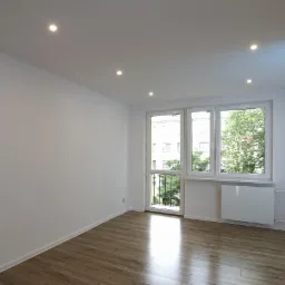Jasne, puste pomieszczenie po remoncie z panelami podłogowymi, białymi ścianami, oknami z widokiem na zieleń i zamontowanymi punktowymi lampami sufitowymi.