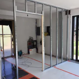 Wnętrze w trakcie budowy ścian działowych z metalowych profili, widoczne instalacje elektryczne i hydrauliczne, podłoga zabezpieczona folią, na zewnątrz widok na ogr&oacute;d przez duże okno.