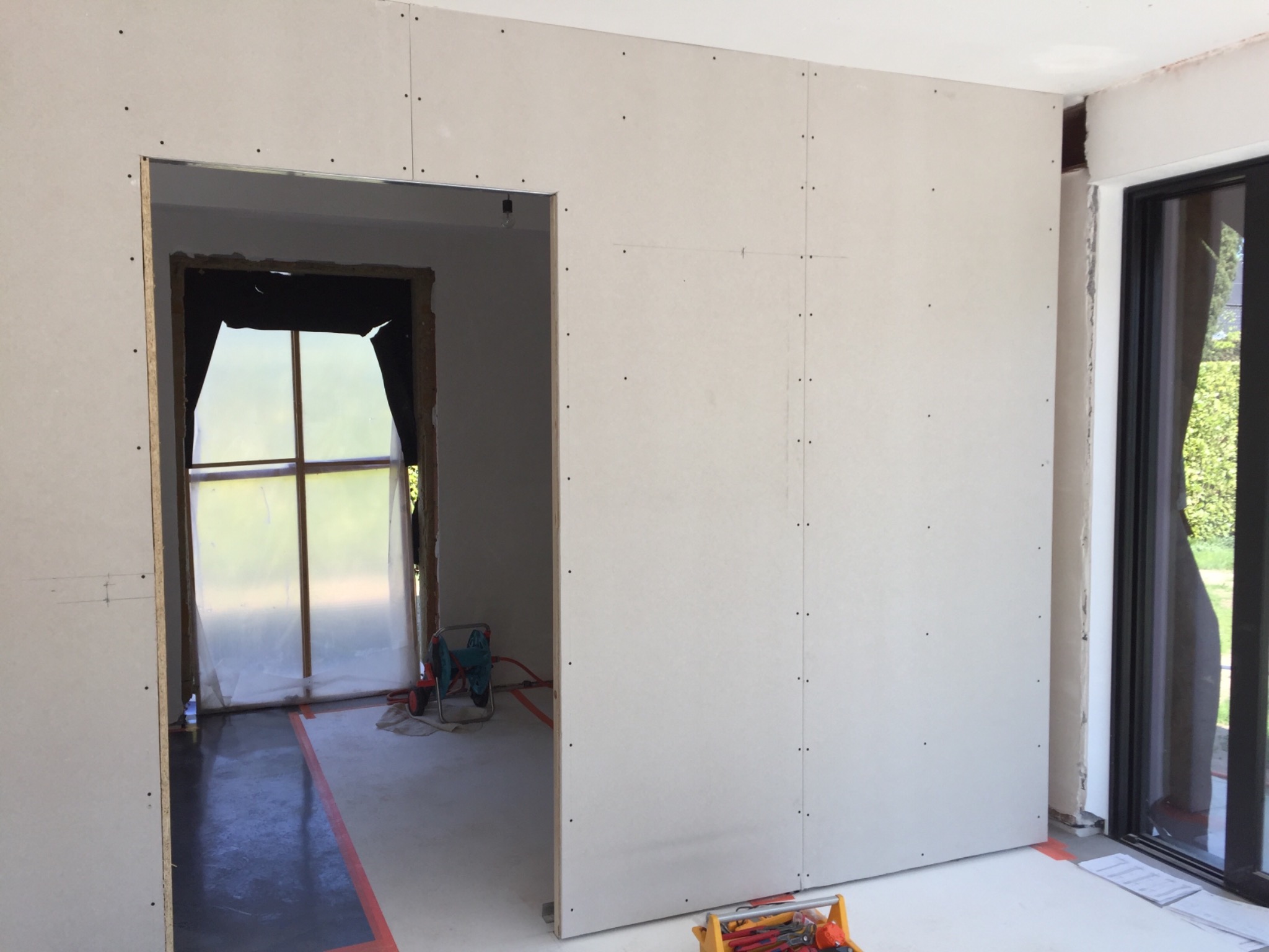 Montaż płyt gipsowo-kartonowych na ścianie, widoczne łączenia płyt, przygotowane otwory drzwiowe i okienne, narzędzia w kuwecie na podłodze, folia ochronna na oknie w tle.