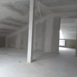 Poddasze w trakcie wykańczania płytami kartonowo-gipsowymi, widoczny słup konstrukcyjny i otwór drzwiowy, ściany przygotowane do dalszych prac wykończeniowych.