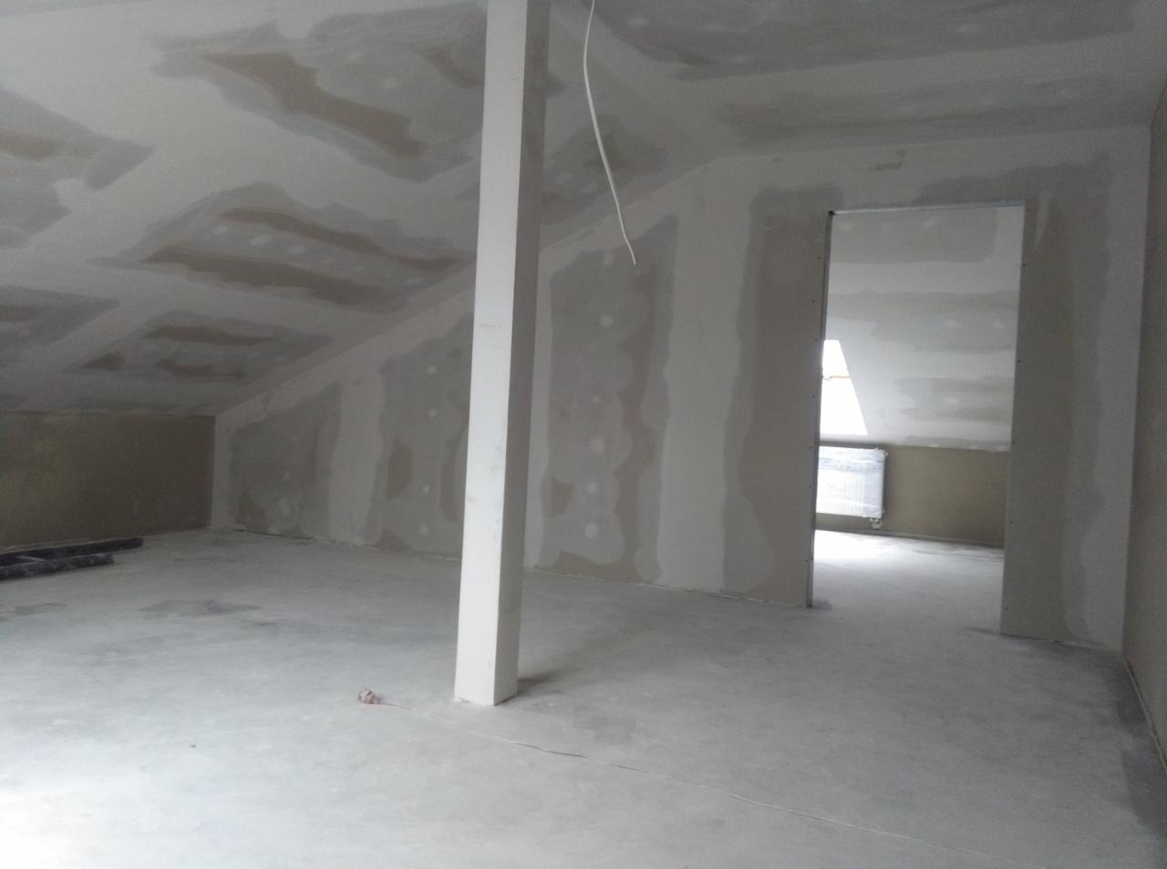 Poddasze w trakcie wykańczania płytami kartonowo-gipsowymi, widoczny słup konstrukcyjny i otwór drzwiowy, ściany przygotowane do dalszych prac wykończeniowych.