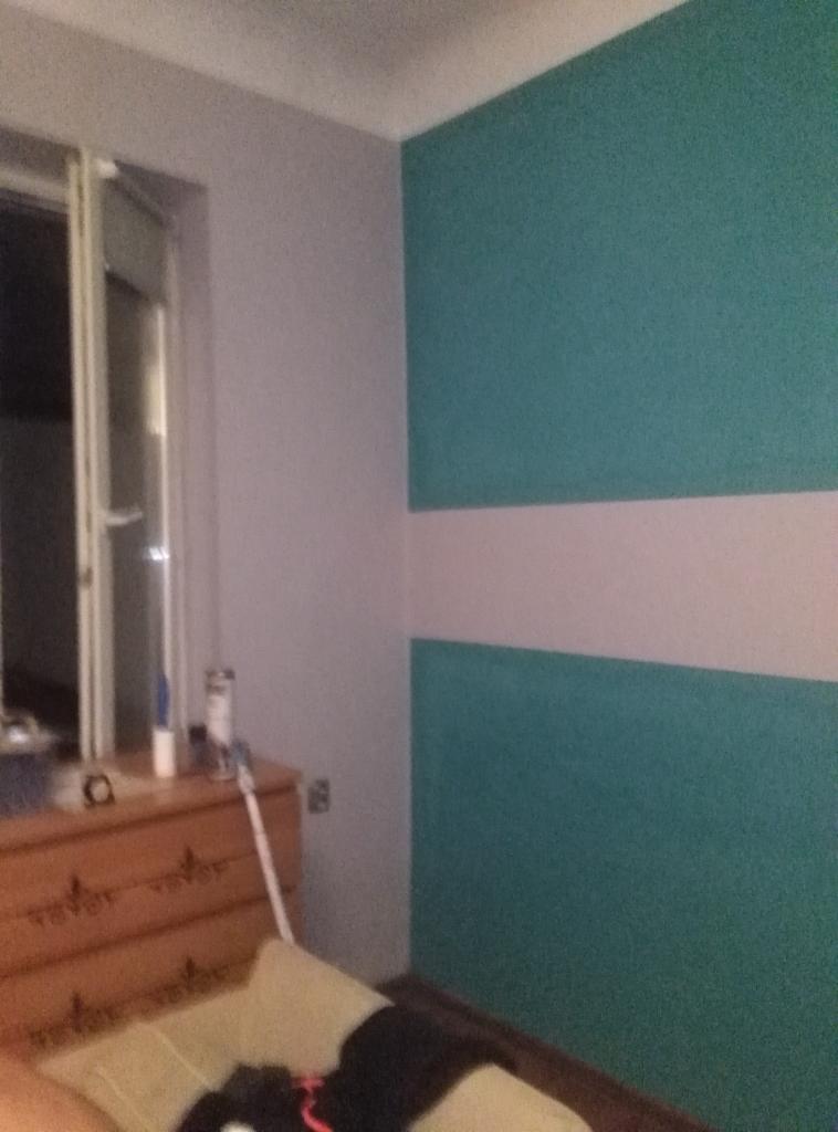 Pomieszczenie z pomalowaną ścianą w poziome pasy turkusu i bieli, widoczna komoda z akcesoriami i fragment łóżka.