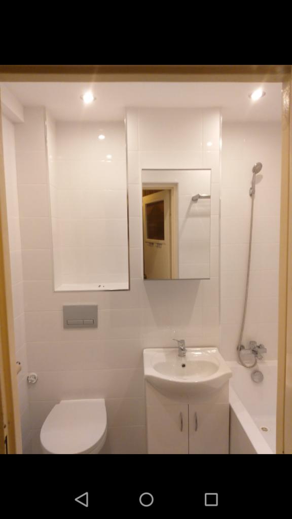 Biała łazienka z umywalką, toaletą, wanną i lustrem, wykończona białymi płytkami, widok na drzwi odbijający się w lustrze.