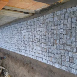 Świeżo ułożona ściana z szarej kostki granitowej, widoczna spod drewnianej konstrukcji stropu, z elementami piasku i ziemi u podstawy.