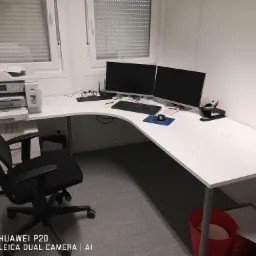 Białe, narożne biurko z dwoma monitorami, klawiaturą, myszką, drukarką, krzesłem obrotowym i koszem na śmieci w rogu pokoju z oknami z żaluzjami.