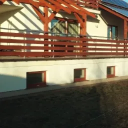 Dom z drewnianą konstrukcją werandy i balkonem, panele słoneczne na dachu, widok z poziomu trawnika.