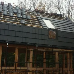 Montaż dachówki ceramicznej w kolorze grafitowym na drewnianej konstrukcji altany z oknem dachowym, widoczne nieułożone dachówki i drzewa w tle.