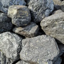 Kupa czarnych brył węgla o zróżnicowanej fakturze, niektóre z połyskiem, inne matowe, oświetlone z góry, tworzące chaotyczny stos.