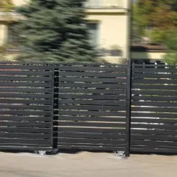 Czarna, pozioma brama wjazdowa z przesuwnymi segmentami na tle rozmytego budynku mieszkalnego i zieleni.