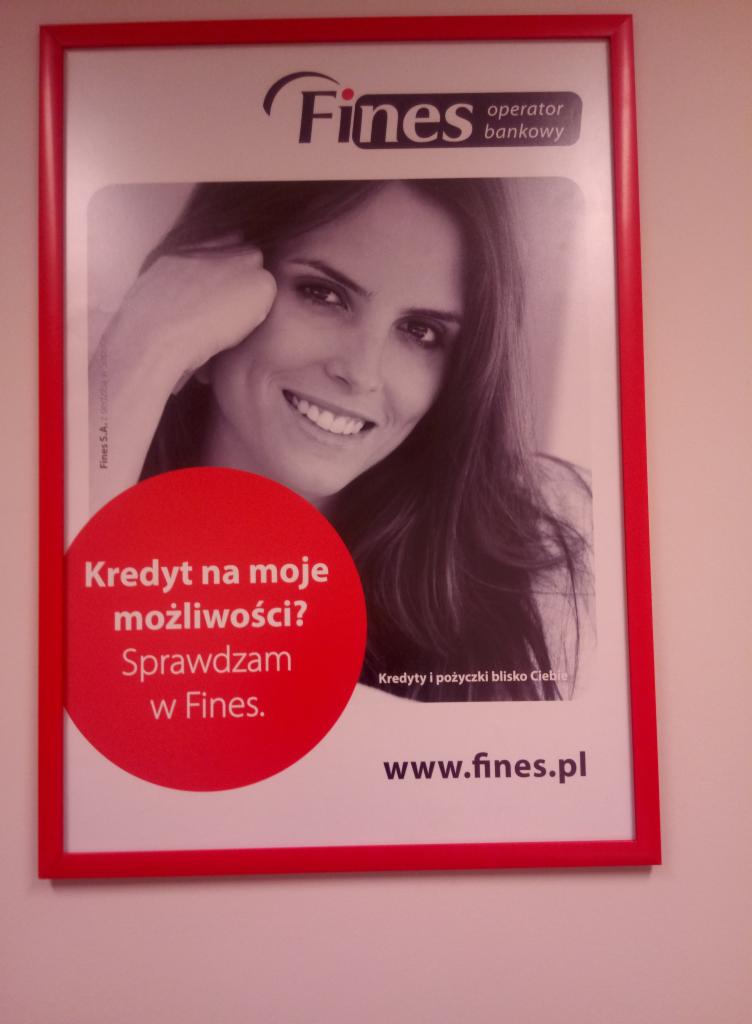 Oprawiony w czerwoną ramkę plakat reklamowy z uśmiechniętą kobietą, logo 'Fines operator bankowy' i hasłem 'Kredyt na moje możliwości? Sprawdzam w Fines.'
