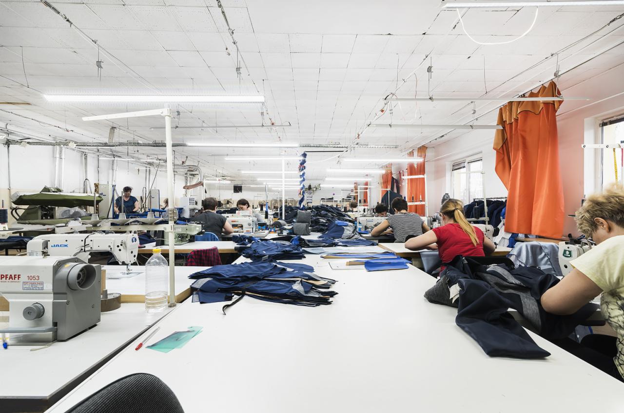 Szewcy pracujący przy maszynach w jasnej szwalni, stosy niebieskich materiałów na stołach, widoczne maszyny Pfaff i Jack.