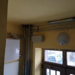 Wewnętrzne piony instalacyjne z rur stalowych i plastikowych, skrzynka elektryczna, biała szafka, oświetlenie sufitowe i okno z widokiem na zaparkowane samochody w pomieszczeniu o żółtych ścianach.