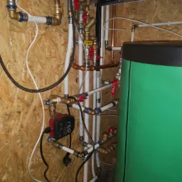 Węzeł cieplny w kotłowni z piecem gazowym, pompą obiegową RCO i zielonym zasobnikiem ciepłej wody użytkowej, widoczne liczne rury miedziane i z tworzywa sztucznego oraz zawory.