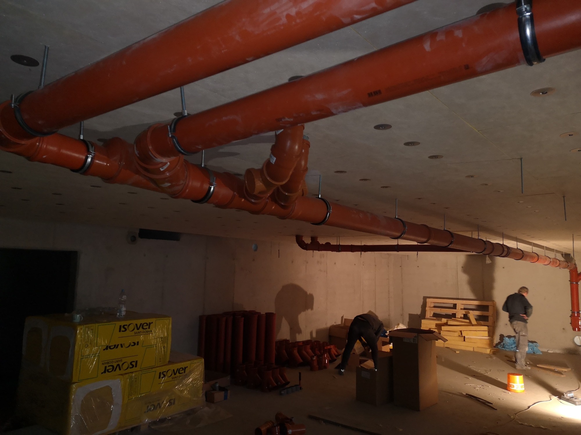 Montaż instalacji kanalizacyjnej: Pomarańczowe rury kanalizacyjne podwieszone pod betonowym sufitem, widoczne złącza i mocowania, w tle pracownicy przy pracy, materiały budowlane składowane...