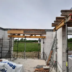 Wznoszenie ścian domu z bloczków Solbet, widoczne podpory i szalunki nad otworami okiennymi, w tle zielony krajobraz.