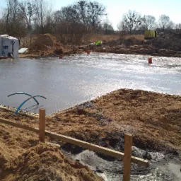 Wylany, mokry fundament pod budynek z widocznymi paletami materiałów SOLBET w tle, przygotowany do dalszych prac.