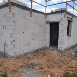 Wznoszenie ścian domu z betonu komórkowego, widoczne zabezpieczenia na górze muru, teren wokół w stanie surowym, widoczny fragment instalacji.