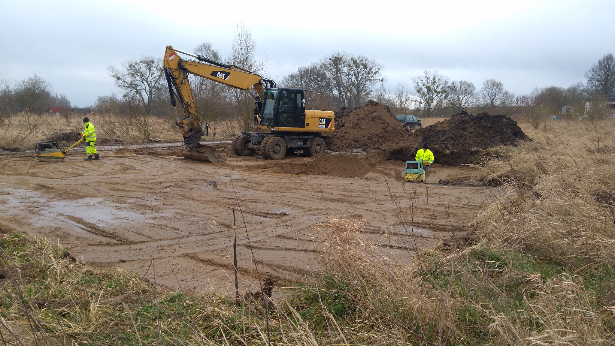 Przygotowanie terenu pod budowę: żółta koparka CAT, ubijarka i dwaj pracownicy w odblaskowych ubraniach na placu budowy z błotnistym podłożem i usypanymi pryzmami ziemi.