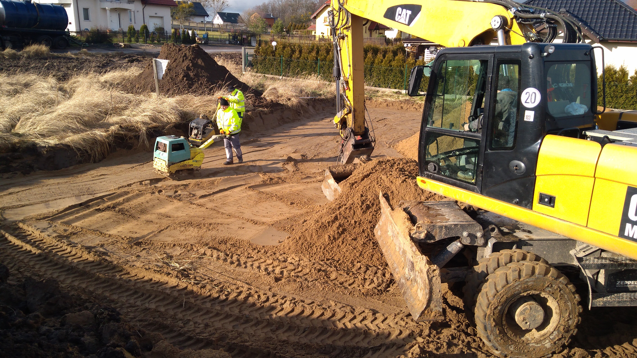 Żółta koparka CAT i zagęszczarka gruntu na placu budowy, dwóch pracowników w kamizelkach odblaskowych, widoczne ślady opon i wykop pod fundamenty.