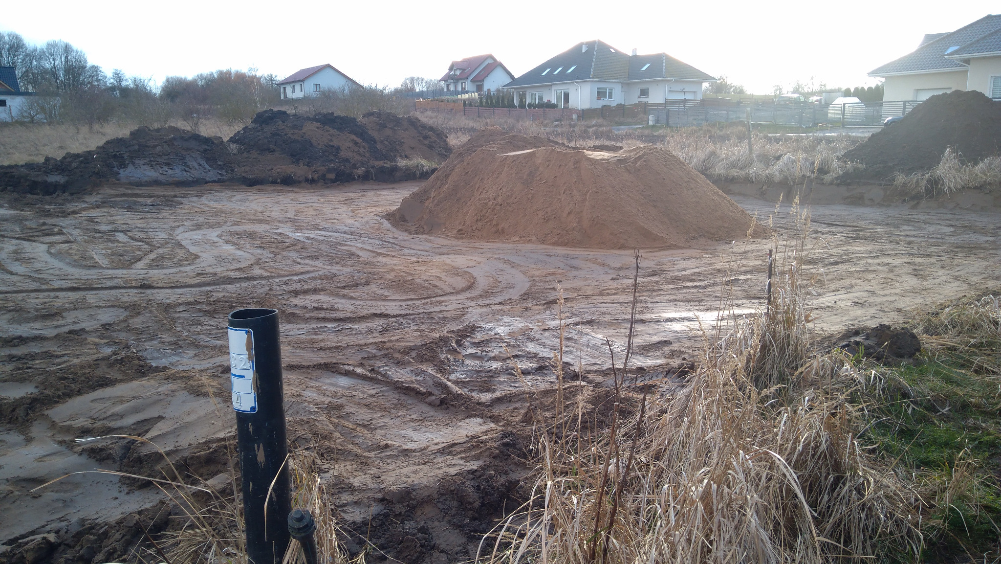 Teren budowy z hałdami piasku i ziemi, widoczne ślady opon, w tle domy jednorodzinne, rura kanalizacyjna z oznaczeniami na pierwszym planie.