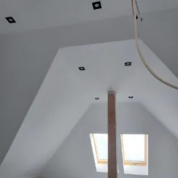 Poddasze po malowaniu na biało, z widocznymi oknami dachowymi i czarnymi, kwadratowymi oprawami oświetleniowymi w suficie, drewniany słup zabezpieczony folią.