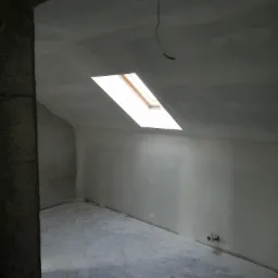 Poddasze w trakcie prac wykończeniowych: ściany i sufit pokryte świeżo nałożoną gładzią, widoczne okno dachowe i wystający kabel elektryczny.