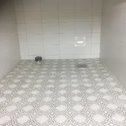 Wykończona kabina prysznicowa z szarymi i białymi płytkami podłogowymi o wzorze marokańskim, białe płytki ścienne, odpływ liniowy w podłodze i czarny element instalacyjny w rogu