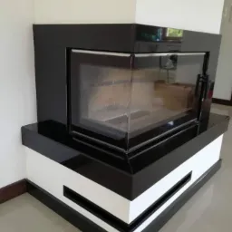 Nowoczesny kominek narożny z czarnego kamienia i białych elementów, widoczny przez szklane drzwiczki paleniska, minimalistyczny design.