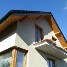 Dom jednorodzinny w trakcie prac elewacyjnych, z widocznym surowym tynkiem, drewnianą podbitką i oknami w drewnianych ramach, balkon w trakcie wykańczania, błękitne niebo w tle.