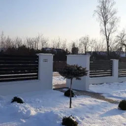 Nowoczesne ogrodzenie panelowe z białymi słupkami i poziomymi, ciemnymi przęsłami, w zimowej scenerii z ośnieżonym trawnikiem i niskimi krzewami.