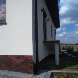 Fragment elewacji domu jednorodzinnego z białym tynkiem i czerwoną cegłą klinkierową u podstawy, widoczne okno z odbiciem chmur i fragment rynny.