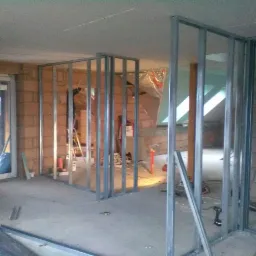 Montaż metalowej konstrukcji ścian działowych w surowym wnętrzu, widoczne profile, cegły, okno dachowe i narzędzia.