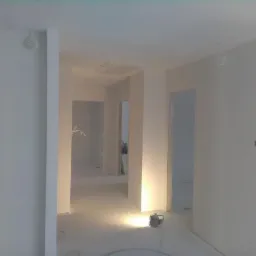 Wnętrze w trakcie remontu, świeżo wykończone ściany w odcieniach bieli, widoczne kable wystające ze ścian, podłoga zabezpieczona folią, lampa budowlana oświetla przestrzeń.